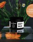 Booty Balm jar with flowers and 'Clean + Conscious Awards' badge on a reflective surface