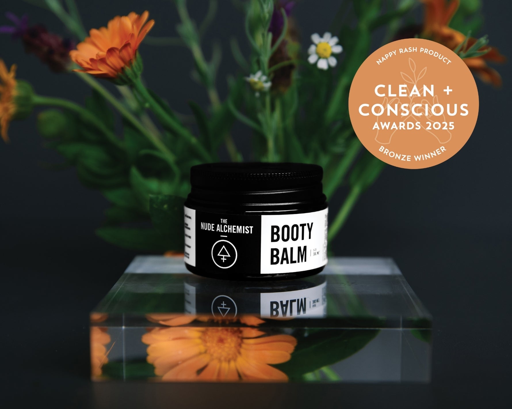 Booty Balm jar with flowers and 'Clean + Conscious Awards' badge on a reflective surface