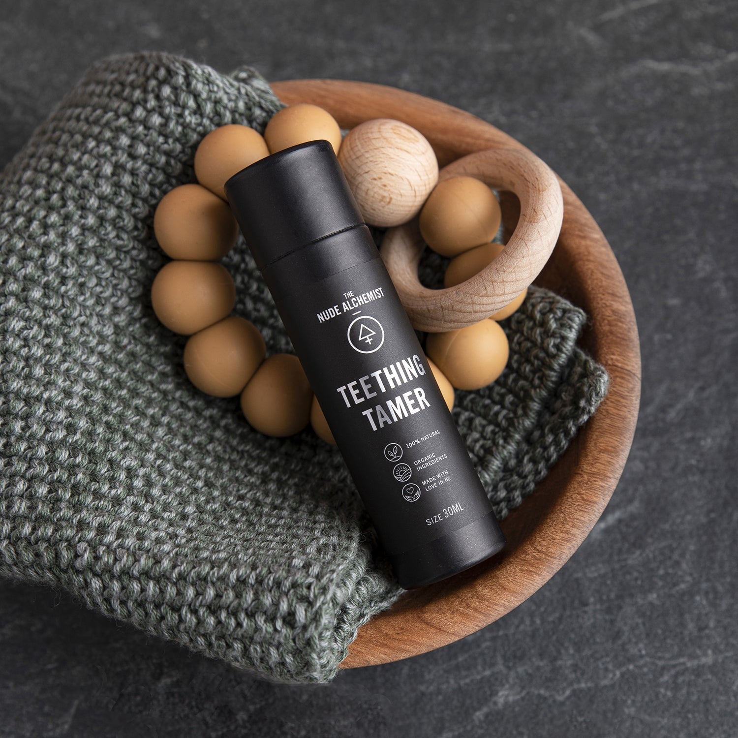 Product shot of Teething Tamer 30g cardboard tube resting on neutral toned teething ring and facecloth in wooden bowl.